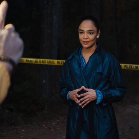 Kvinde i mørk frakke poserer foran et kamera med en politiafspærring bag hende.